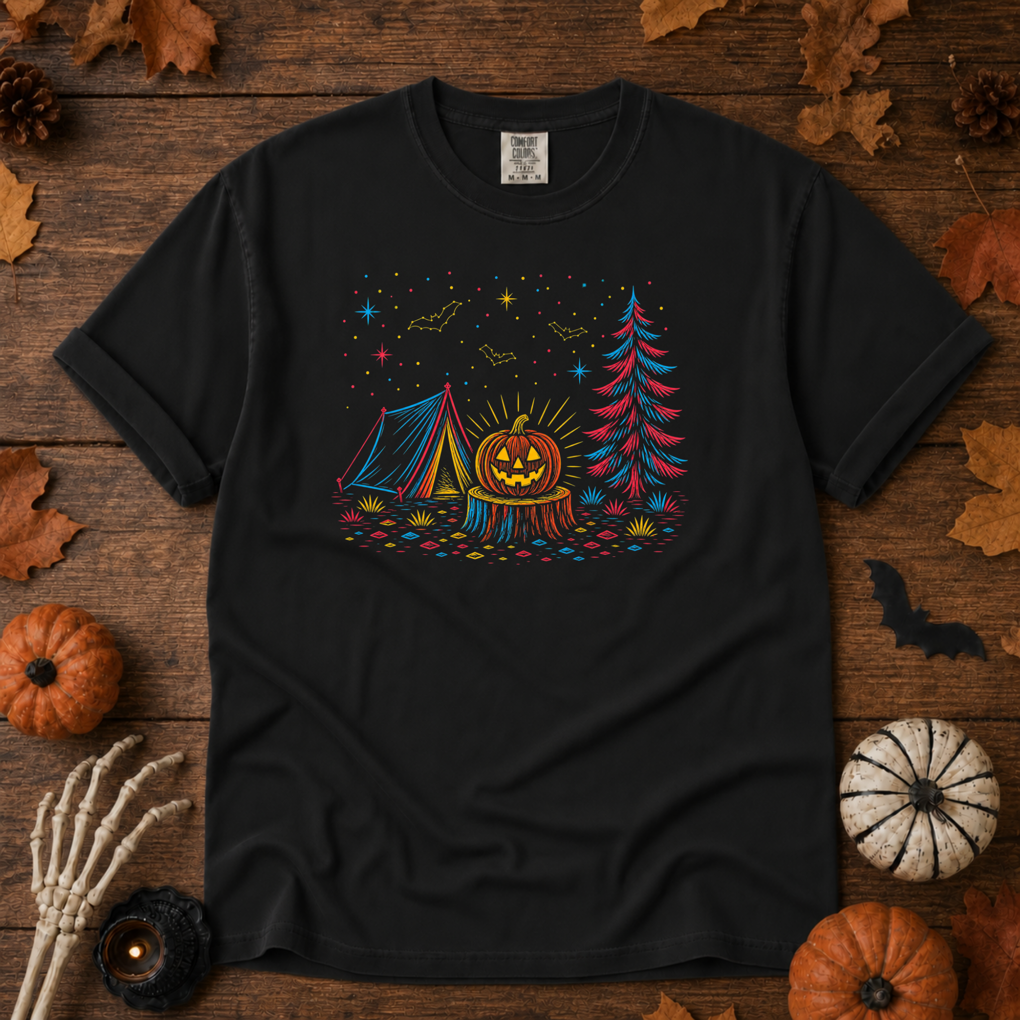 Black Comfort Colors t-shirt featuring a neon-lit Halloween campsite scene — glowing tent, jack-o'-lantern on a stump, pine tree, bats and stars in red, blue, and gold, Trail & Ember Co.