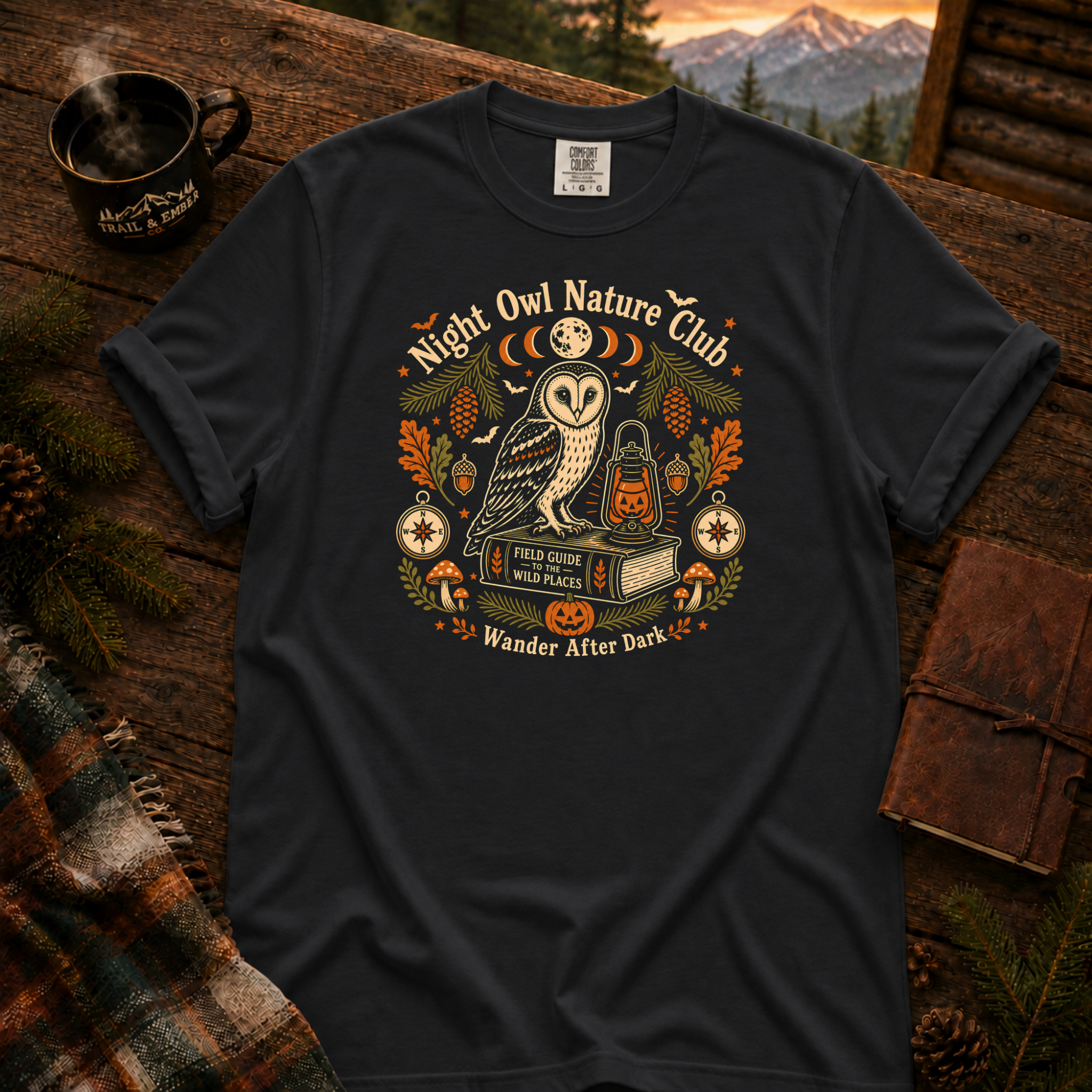 Black Comfort Colors t-shirt featuring a vintage Night Owl Nature Club badge design — barn owl perched on a field guide book beside a jack-o'-lantern lantern, moon phases, compass, pinecones, bats, and autumn leaves, Trail & Ember Co.