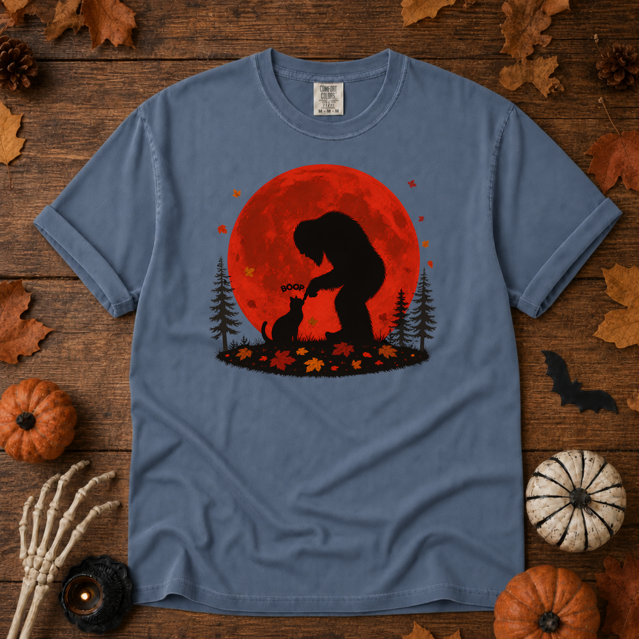 Blue Jean Comfort Colors t-shirt featuring Bigfoot booping a black cat's nose in front of a red autumn moon, with maple leaves and pine trees, Trail & Ember Co.