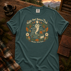 Blue Spruce Comfort Colors t-shirt featuring a vintage Night Owl Nature Club badge design — barn owl perched on a field guide book beside a jack-o'-lantern lantern, moon phases, compass, pinecones, bats, and autumn leaves, Trail & Ember Co.