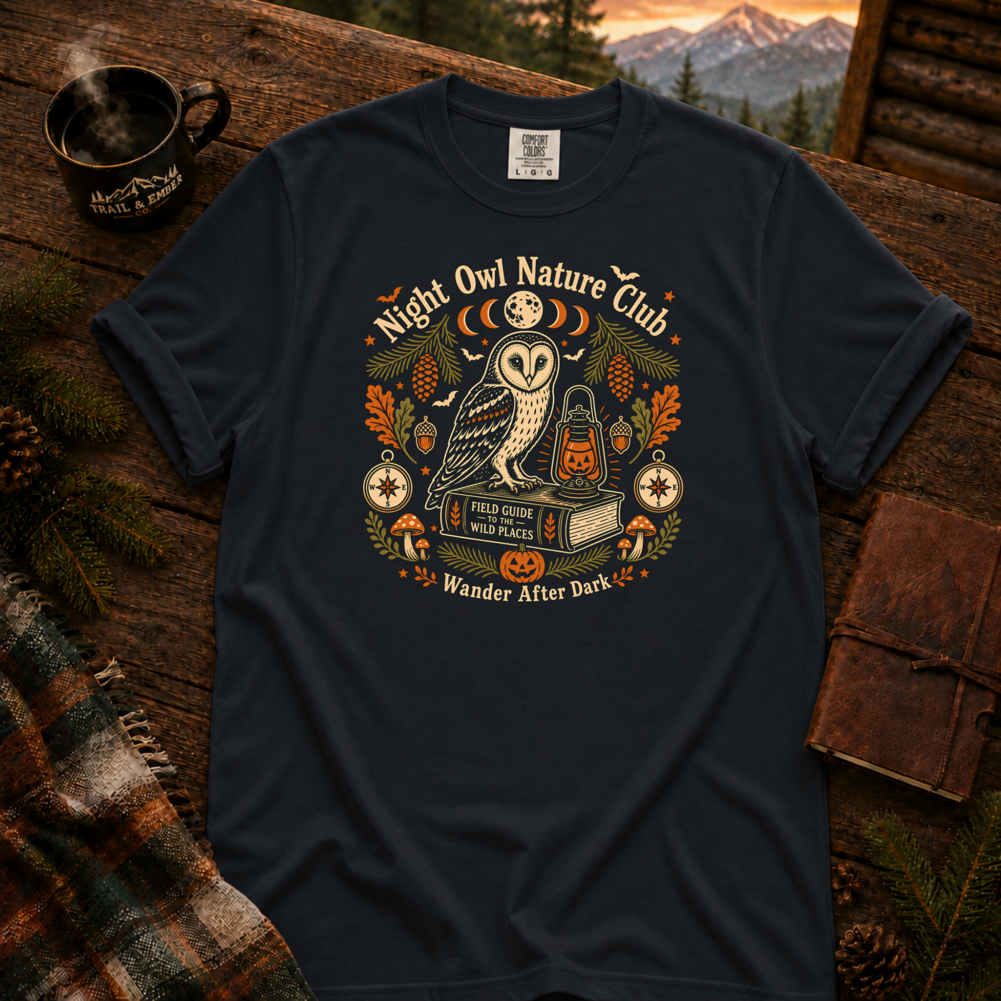 Graphite Comfort Colors t-shirt featuring a vintage Night Owl Nature Club badge design — barn owl perched on a field guide book beside a jack-o'-lantern lantern, moon phases, compass, pinecones, bats, and autumn leaves, Trail & Ember Co.
