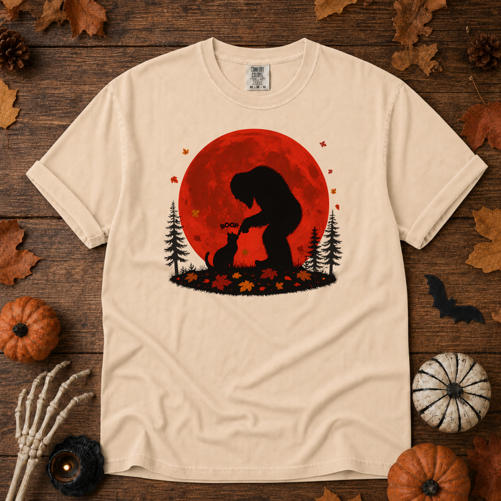 Ivory Comfort Colors t-shirt featuring Bigfoot booping a black cat's nose in front of a red autumn moon, with maple leaves and pine trees, Trail & Ember Co.