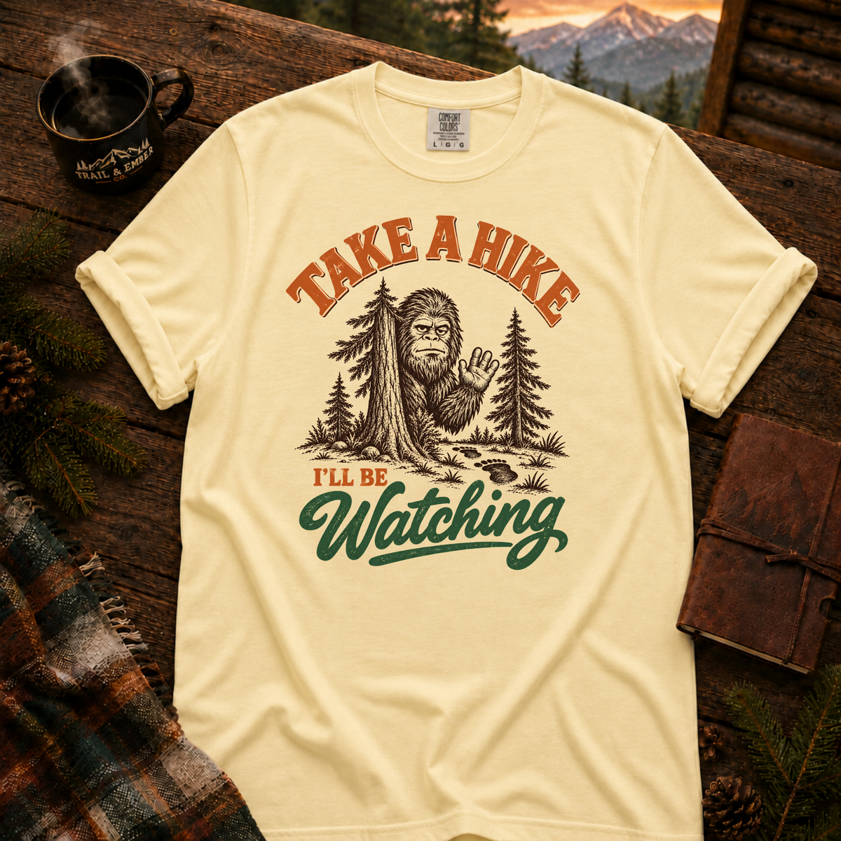 Butter Comfort Colors T-shirt with Take a Hike, I'll Be Watching Bigfoot waving behind a pine tree.