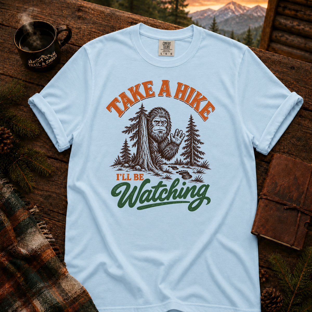 Chambray Comfort Colors T-shirt with Take a Hike, I'll Be Watching Bigfoot waving behind a pine tree.