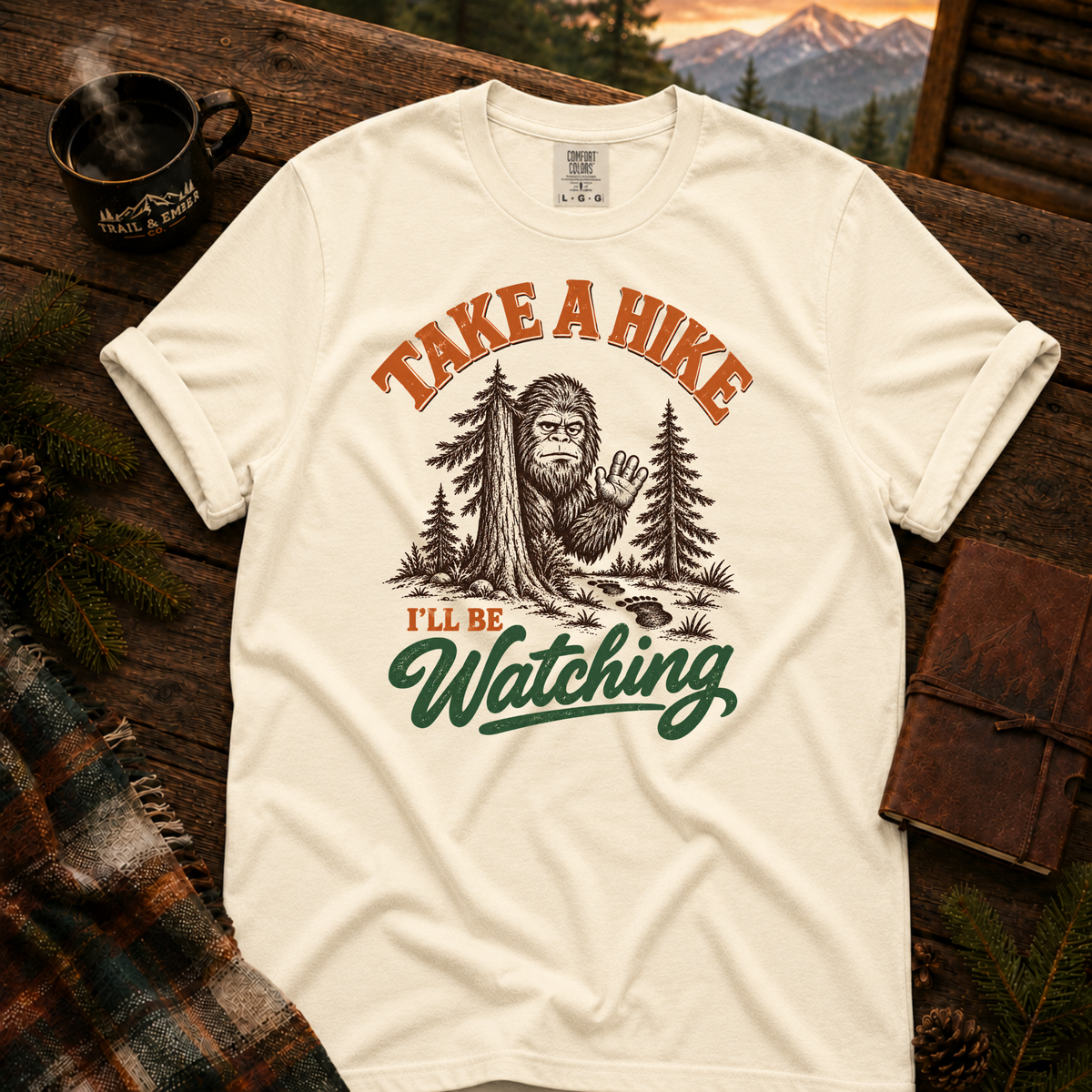 Ivory Comfort Colors T-shirt with Take a Hike, I'll Be Watching Bigfoot waving behind a pine tree.