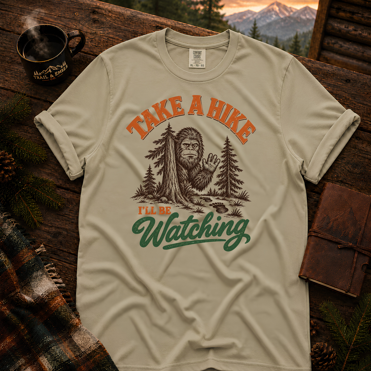 Khaki Comfort Colors T-shirt with Take a Hike, I'll Be Watching Bigfoot waving behind a pine tree.